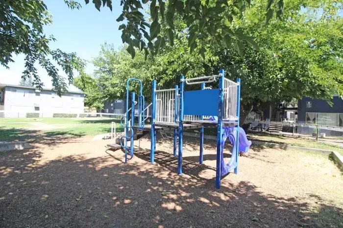 Playground structure in a park, blue and silver, surrounded by wood chips and trees.