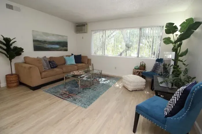 Living room with tan couch, blue rug, and two blue chairs. Bright windows and large plant.
