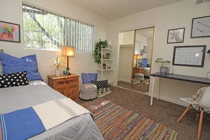 Bedroom with bed, desk, and sliding door to another room. Neutral tones with accents of blue, green, and wood.