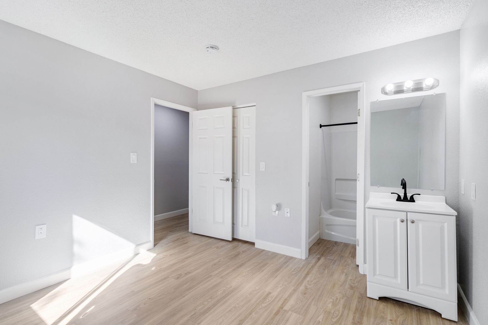 Empty, light-filled bathroom with white vanity, shower, and doors, light wood-look flooring, and gray walls.