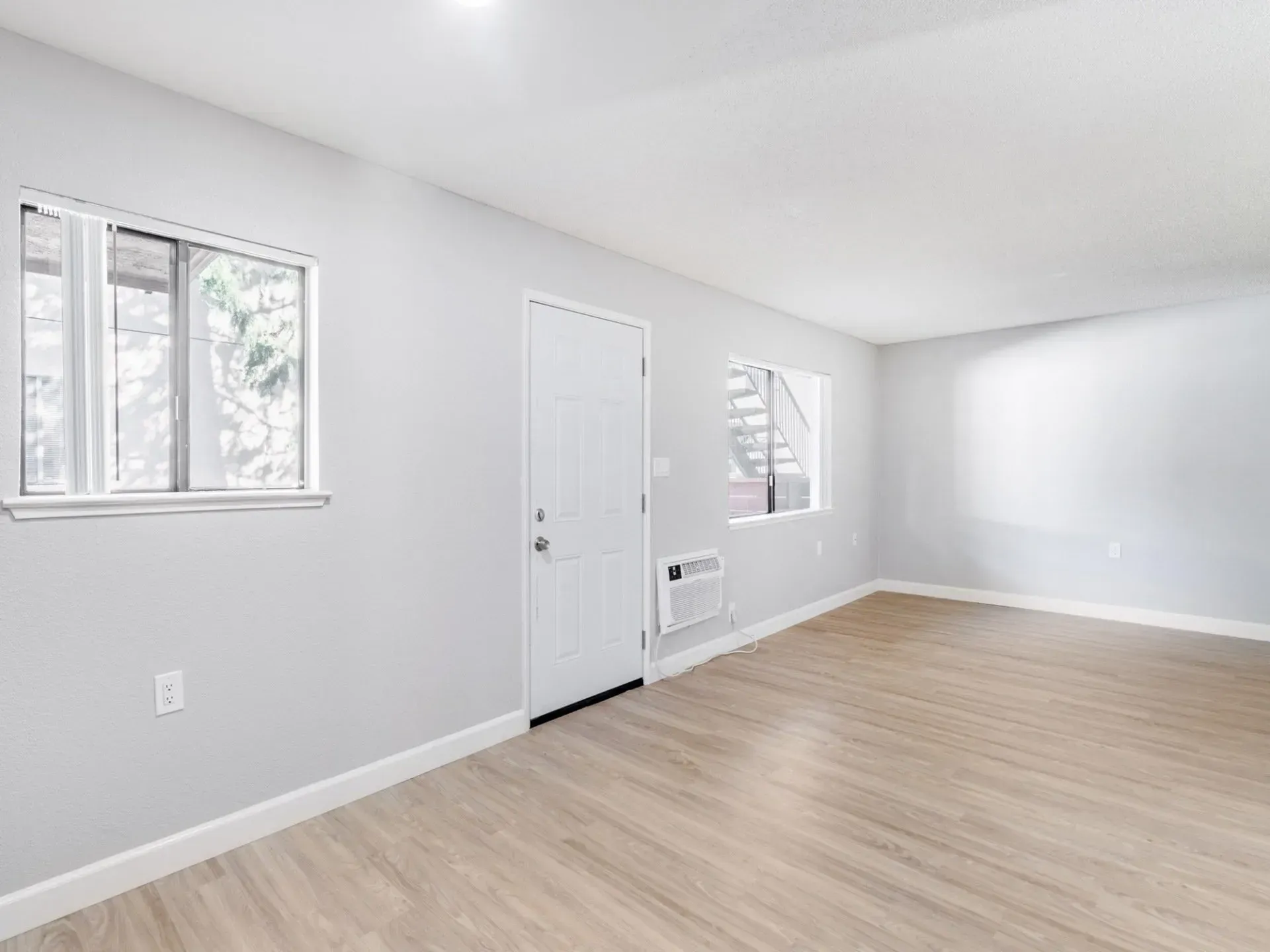 Empty, bright room with white walls, wood-look flooring, door, windows, and an air conditioner.