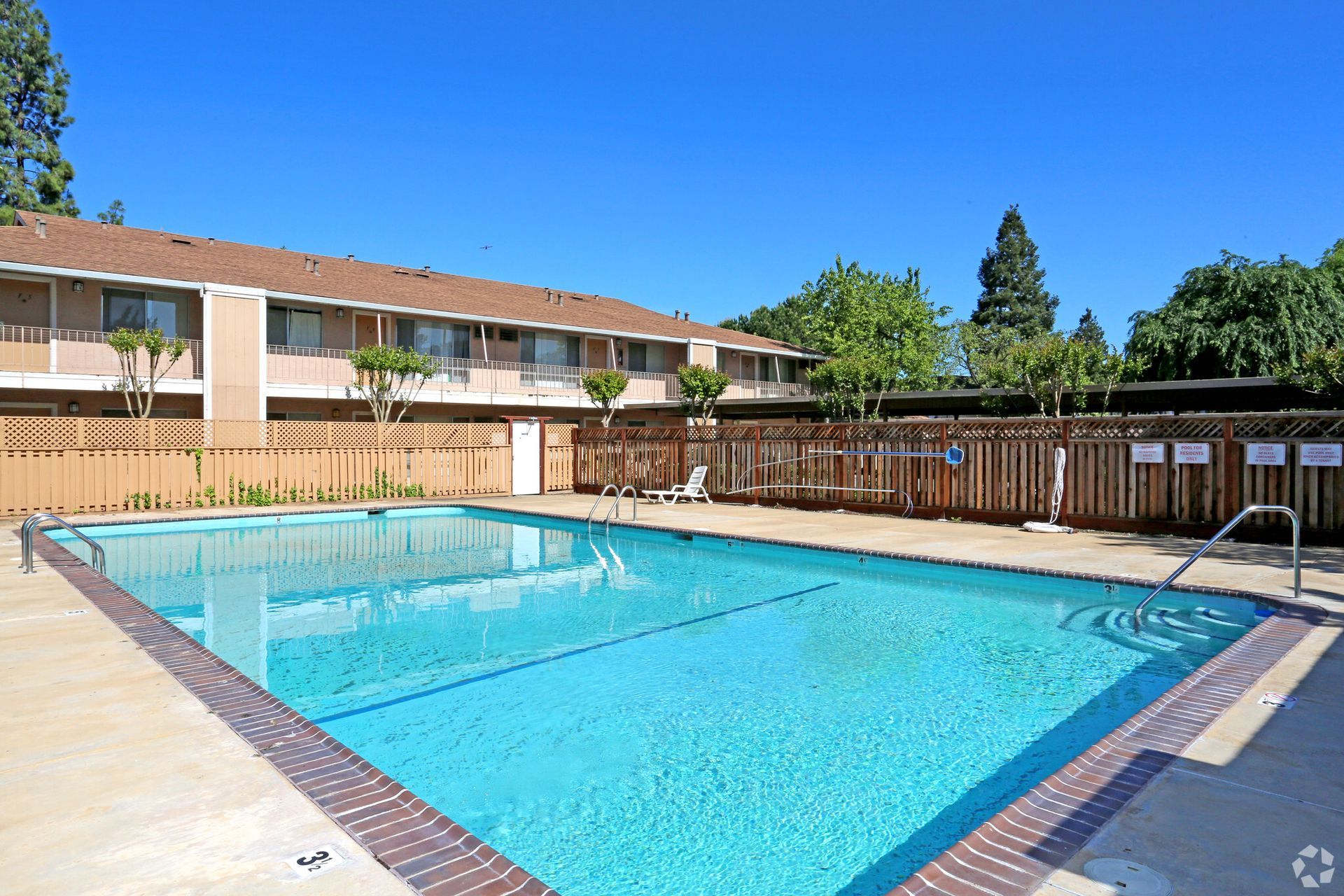 Apartment complex with a rectangular swimming pool on a sunny day. Brown buildings, blue sky.