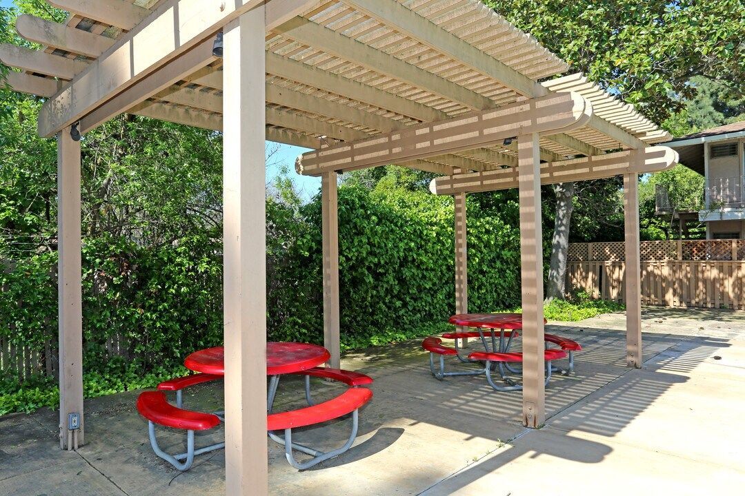 Picnic tables under a wooden pergola shelter. Red tables and benches, surrounded by greenery.