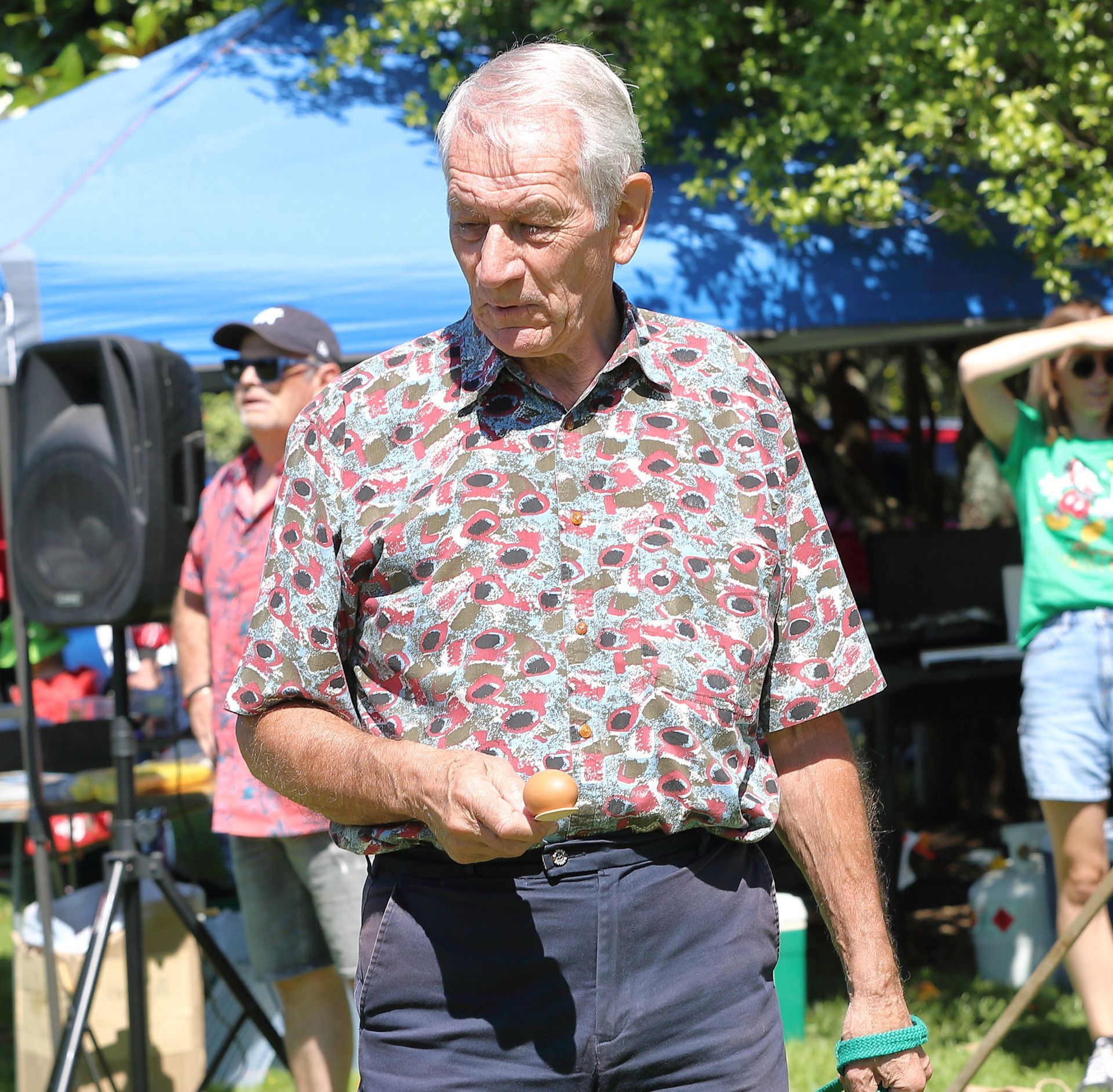Man in patterned shirt holding an egg, outdoors. Tent and people in the background.