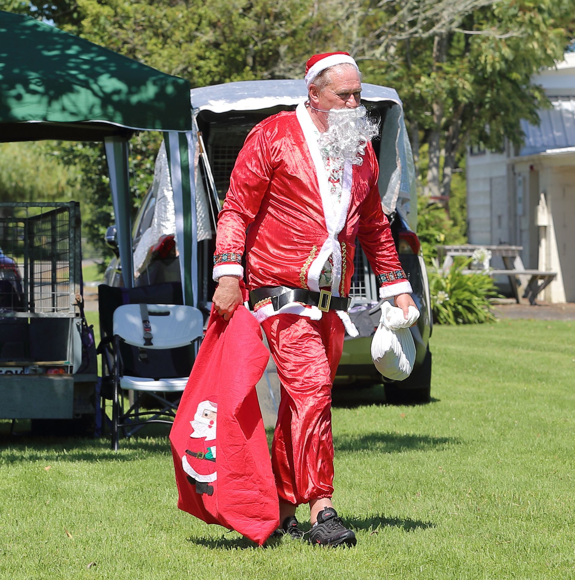 Santa carrying a sack and a white bag, walking on grass near a vehicle.