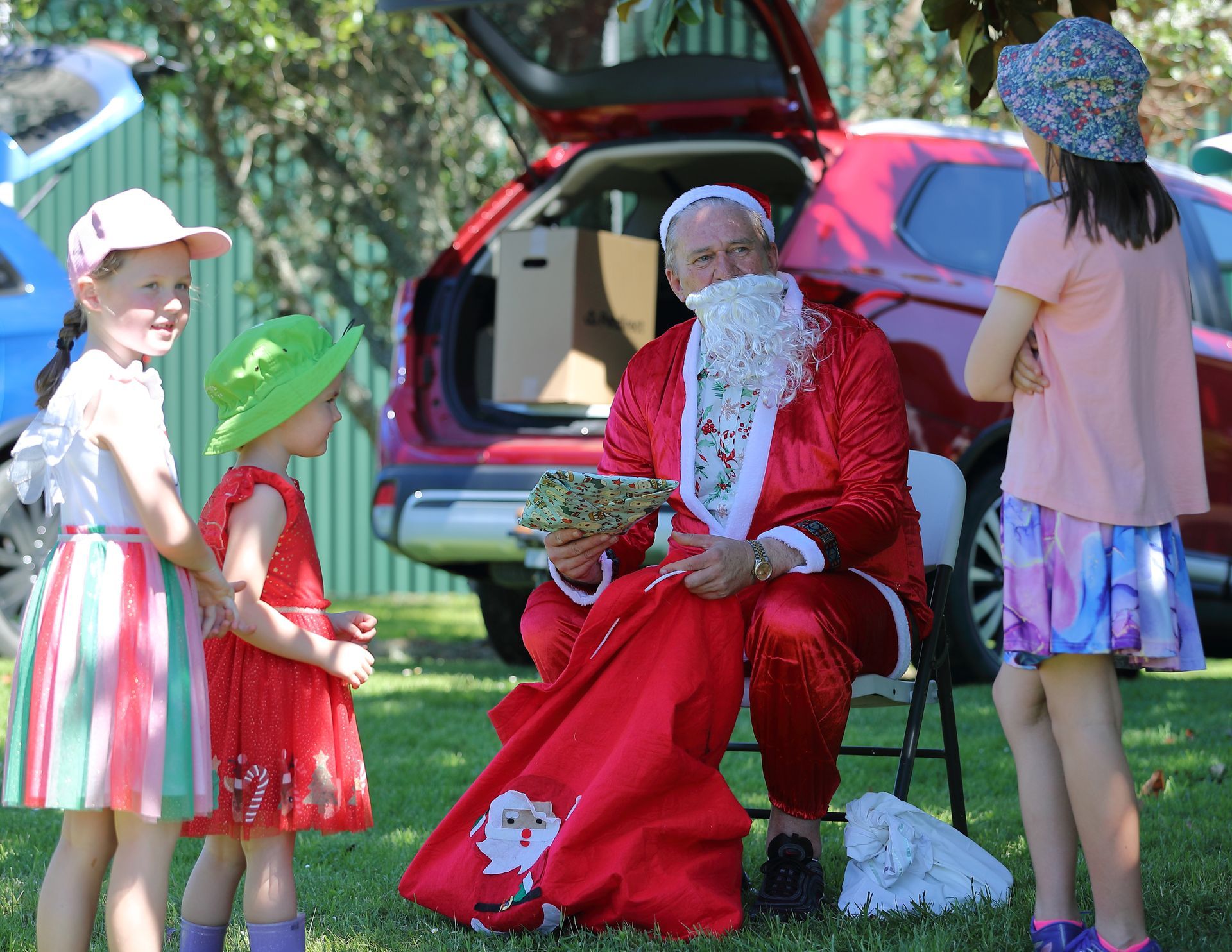 Santa Claus in red suit with children outdoors; Santa holds gift bag near car.