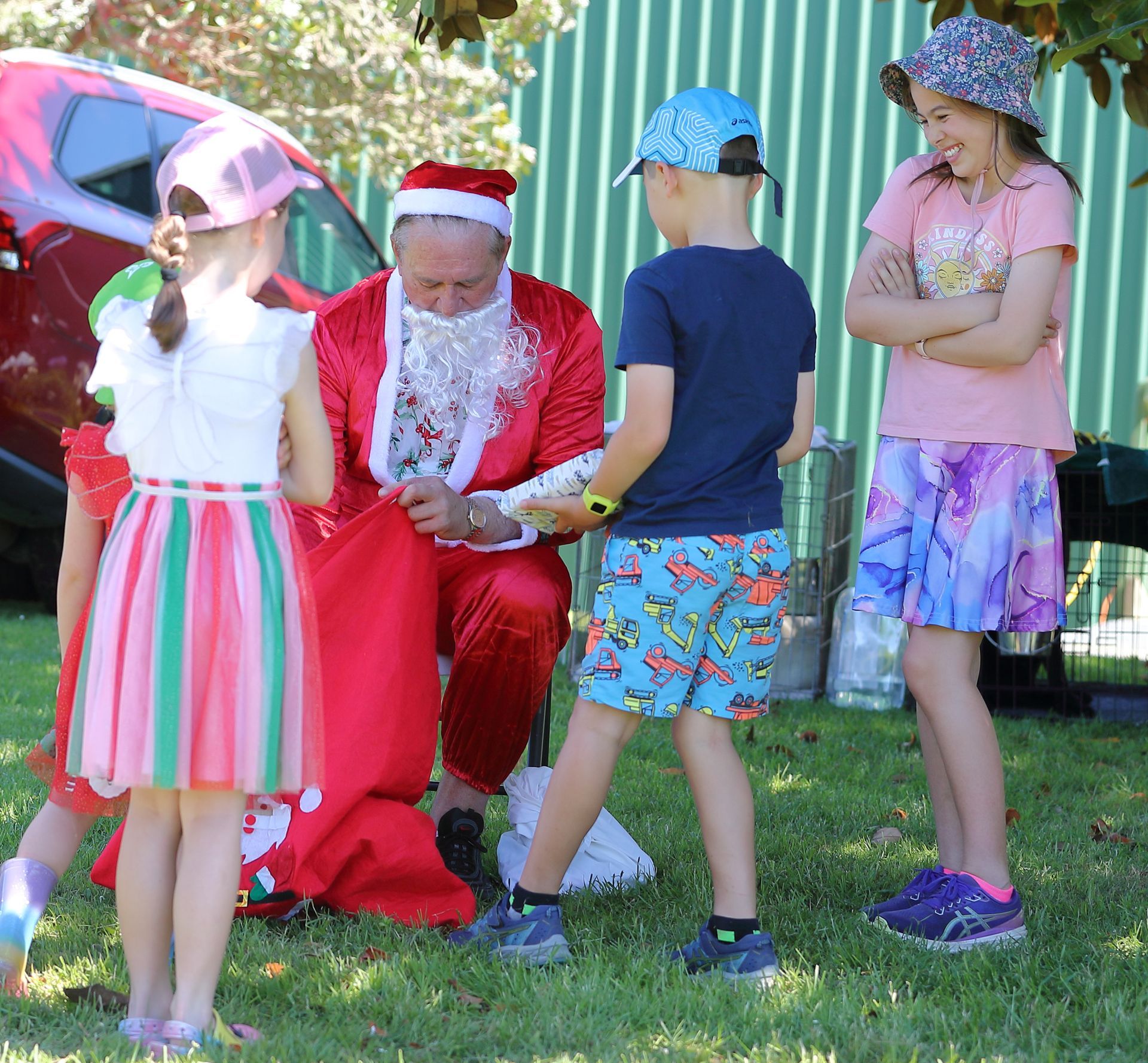 Santa kneels, handing gifts to children outdoors. Colorful clothing, green grass, a red car.