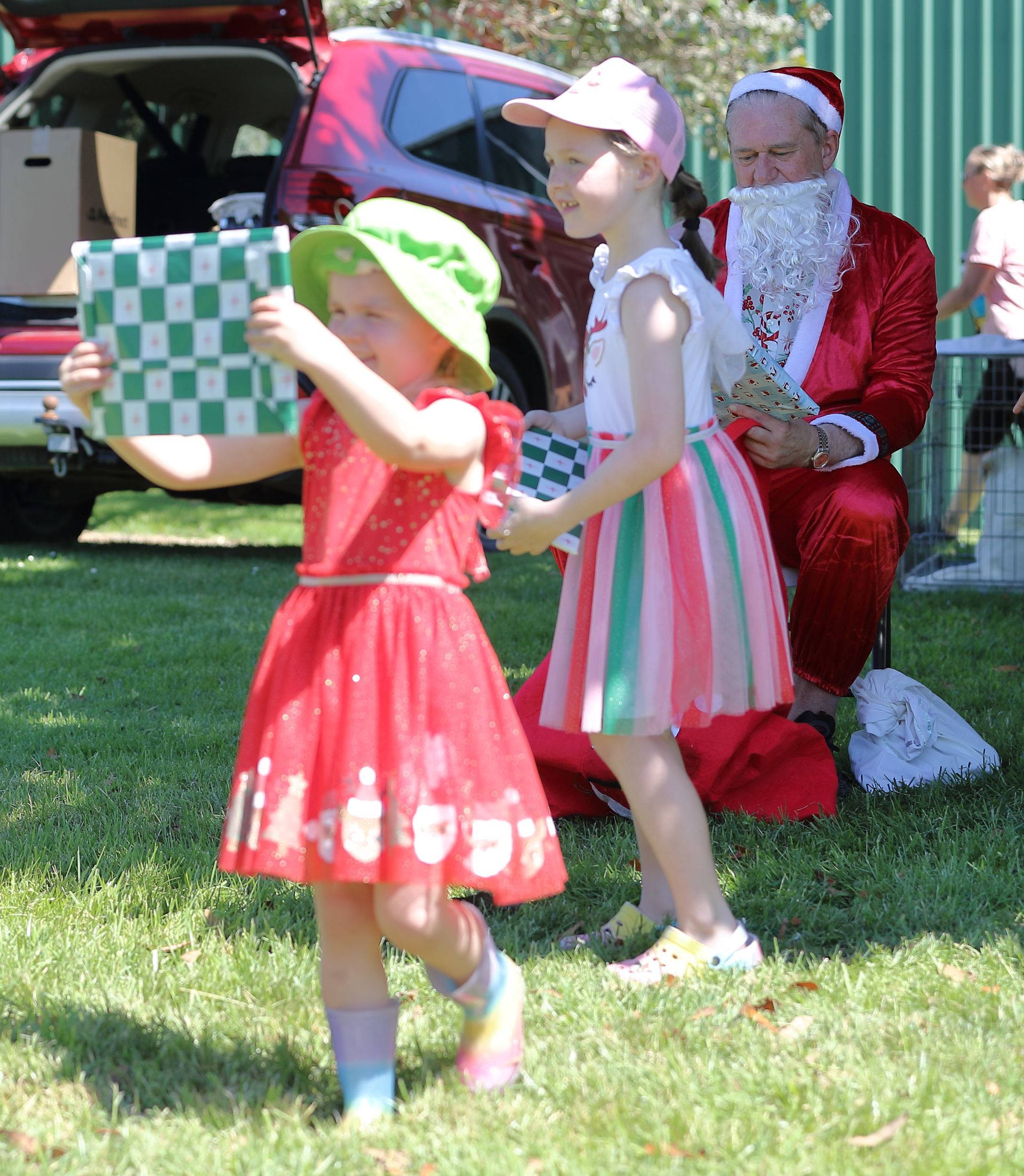 Two girls hold checkerboard gifts, Santa watches outdoors.