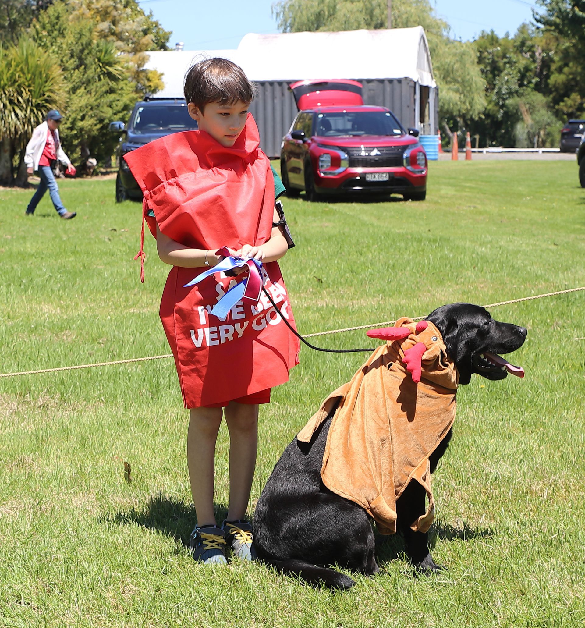 Boy in red costume and black dog in costume on green grass; cars and building in background.