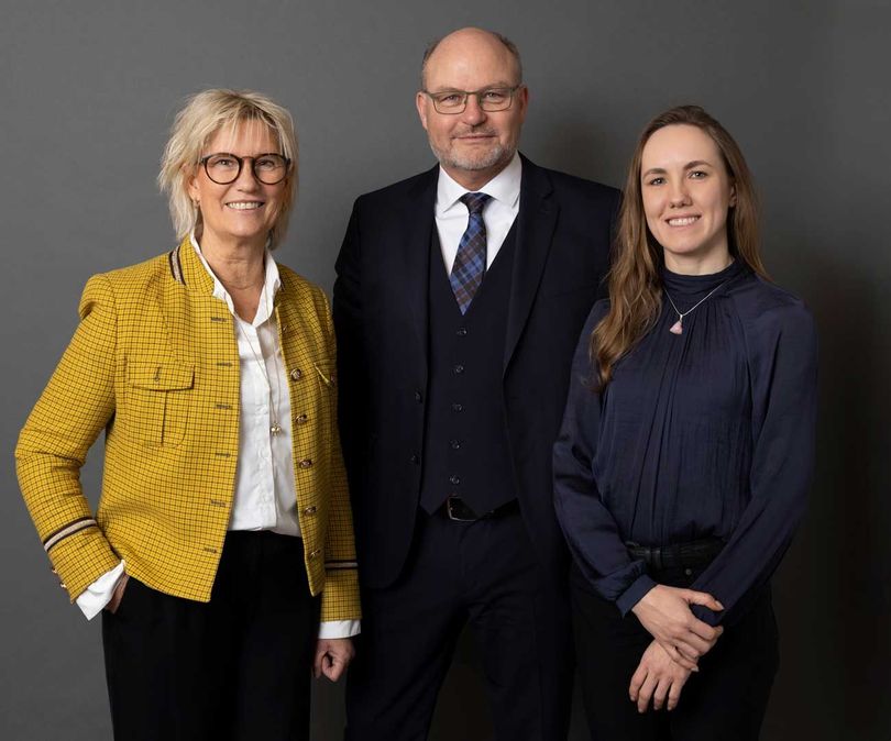 Tre personer poserer foran en grå baggrund: en kvinde i gul jakke, en mand i jakkesæt og en kvinde i blå bluse.