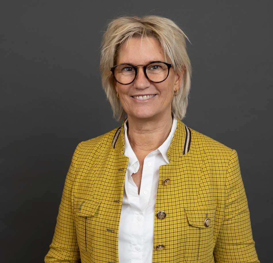En smilende person med blond hår og briller, iført en hvid skjorte og en gulmønstret blazer mod en mørk baggrund.