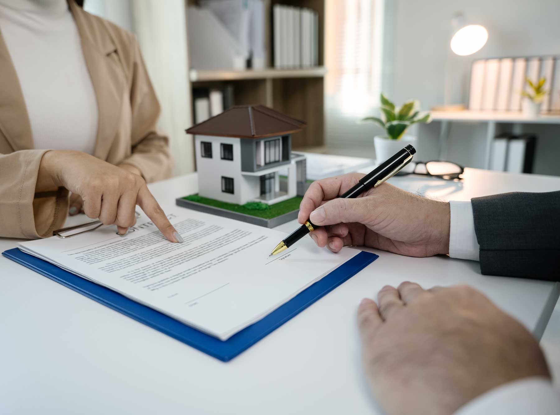 Person peger på et dokument, mens en anden person underskriver det; modelhus på et skrivebord.