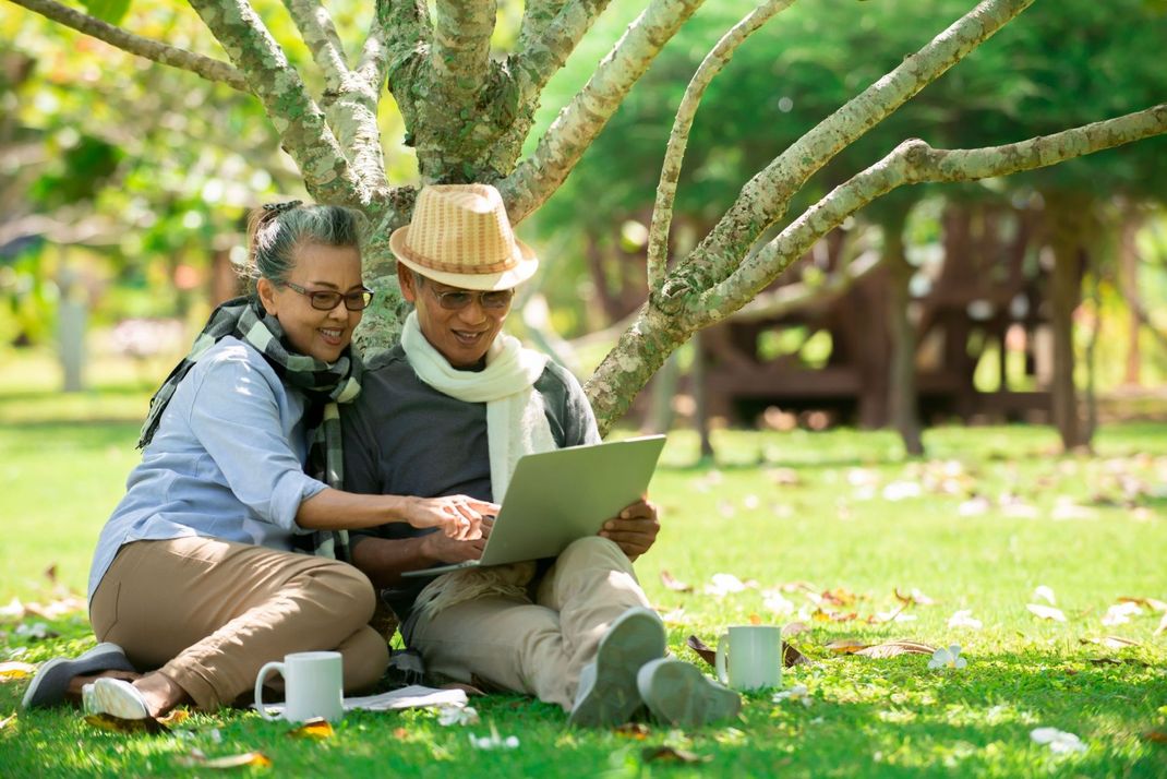 An elderly couple is sitting under a tree looking at a laptop.