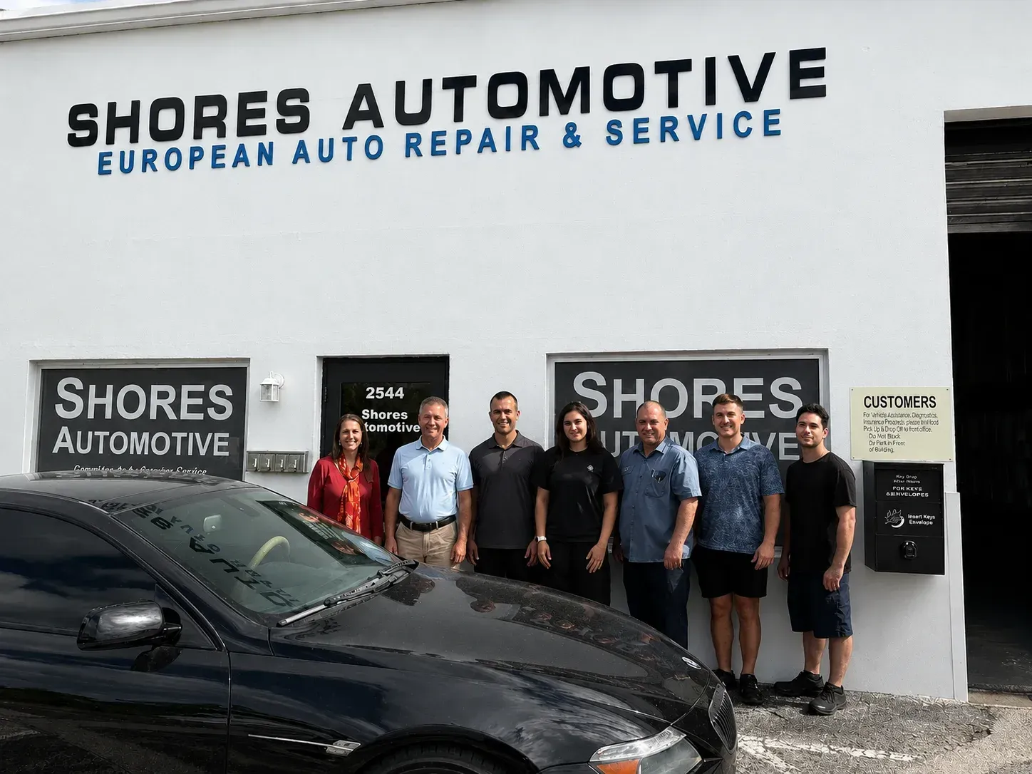 People standing outside Shores Automotive, an auto repair shop, with a black car in front