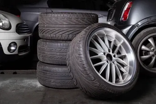 Stacked black tires with silver alloy wheels in a garage, with a car partially visible beside them