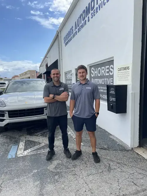Two men standing outside Suores Automotive beside a white SUV on a cloudy day.