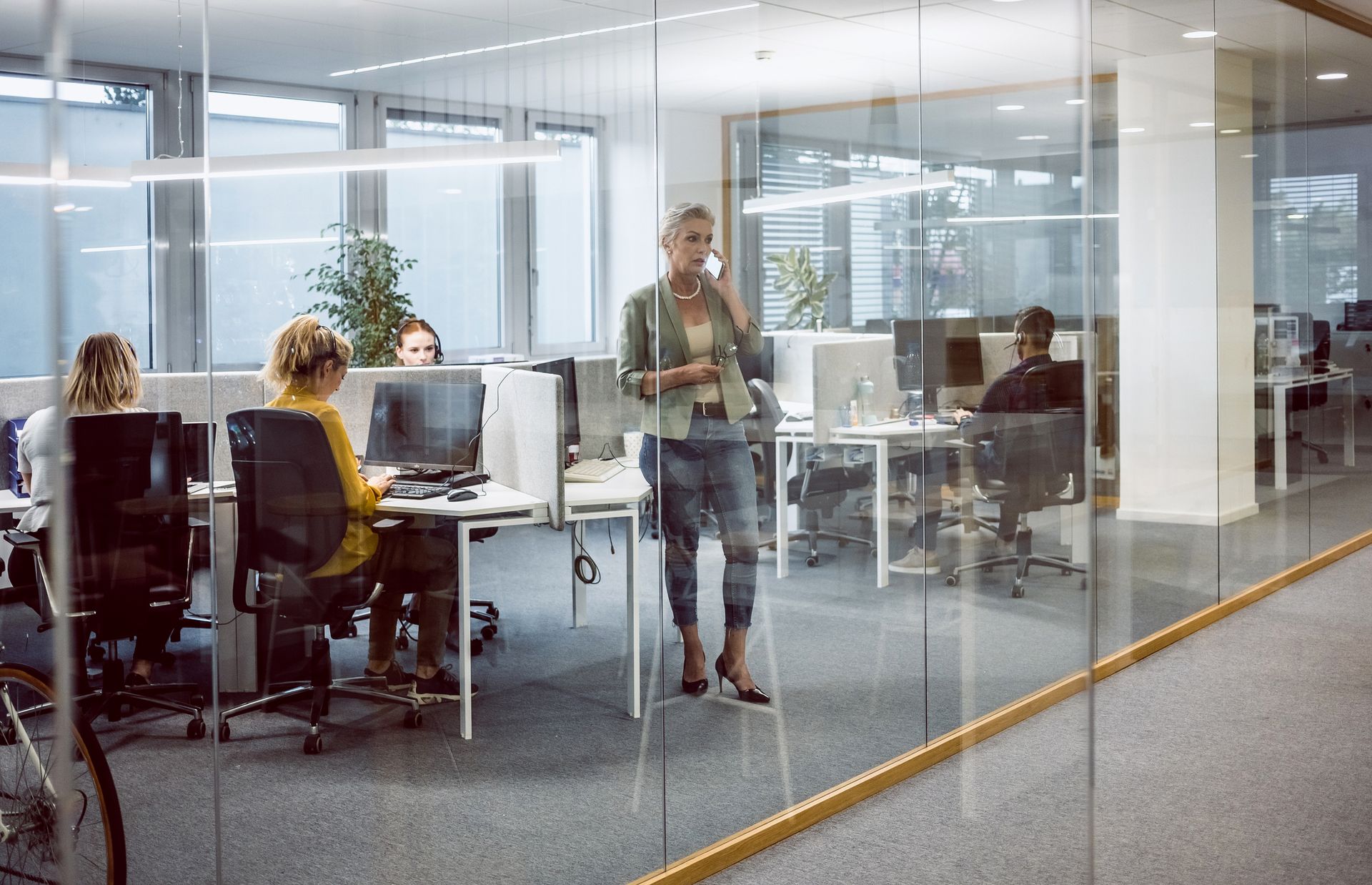 Woman on phone in office, standing near glass-walled workspace with other employees at computers.