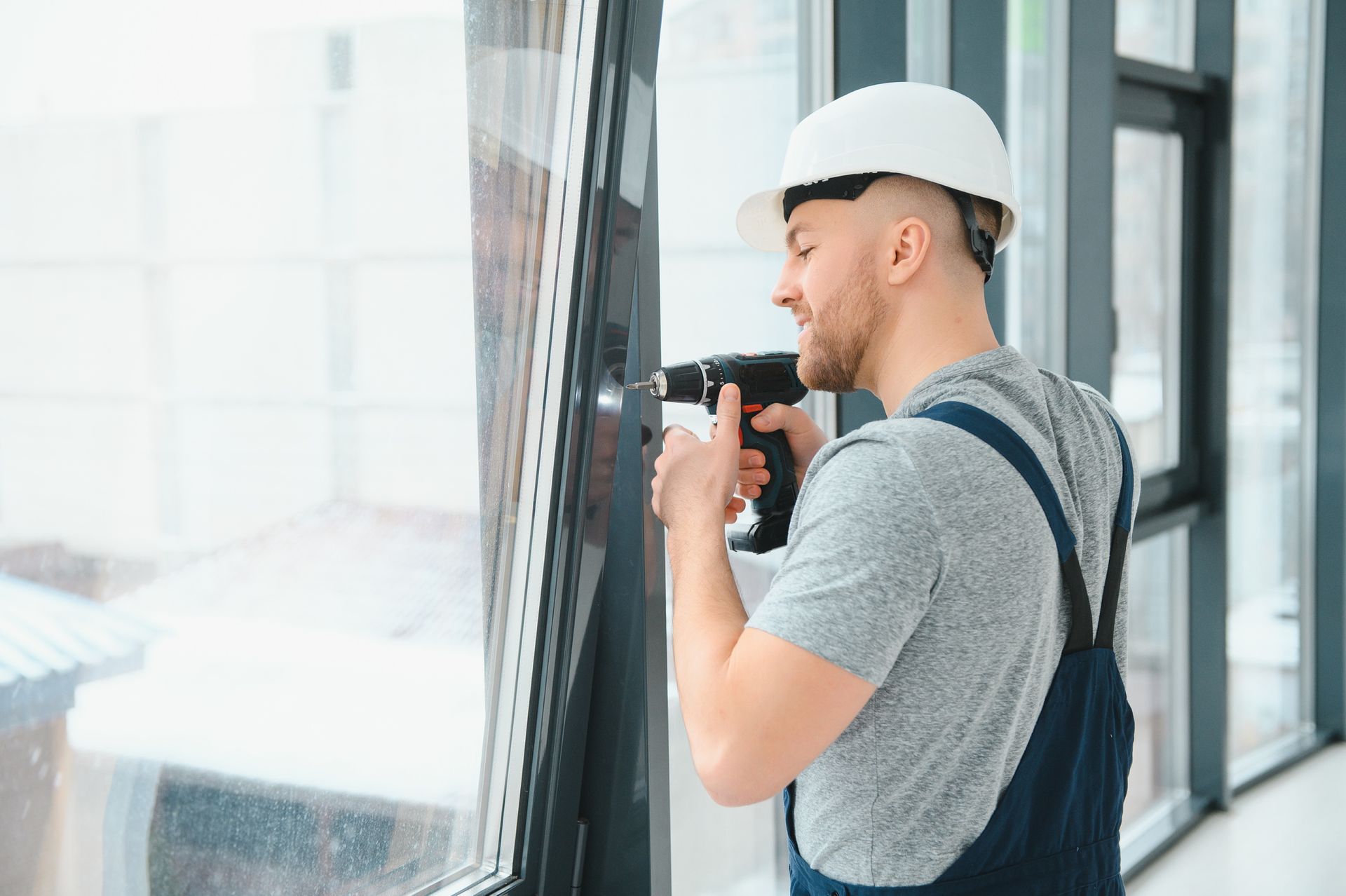 Construction worker drilling a window frame. White hard hat, blue overalls, interior.