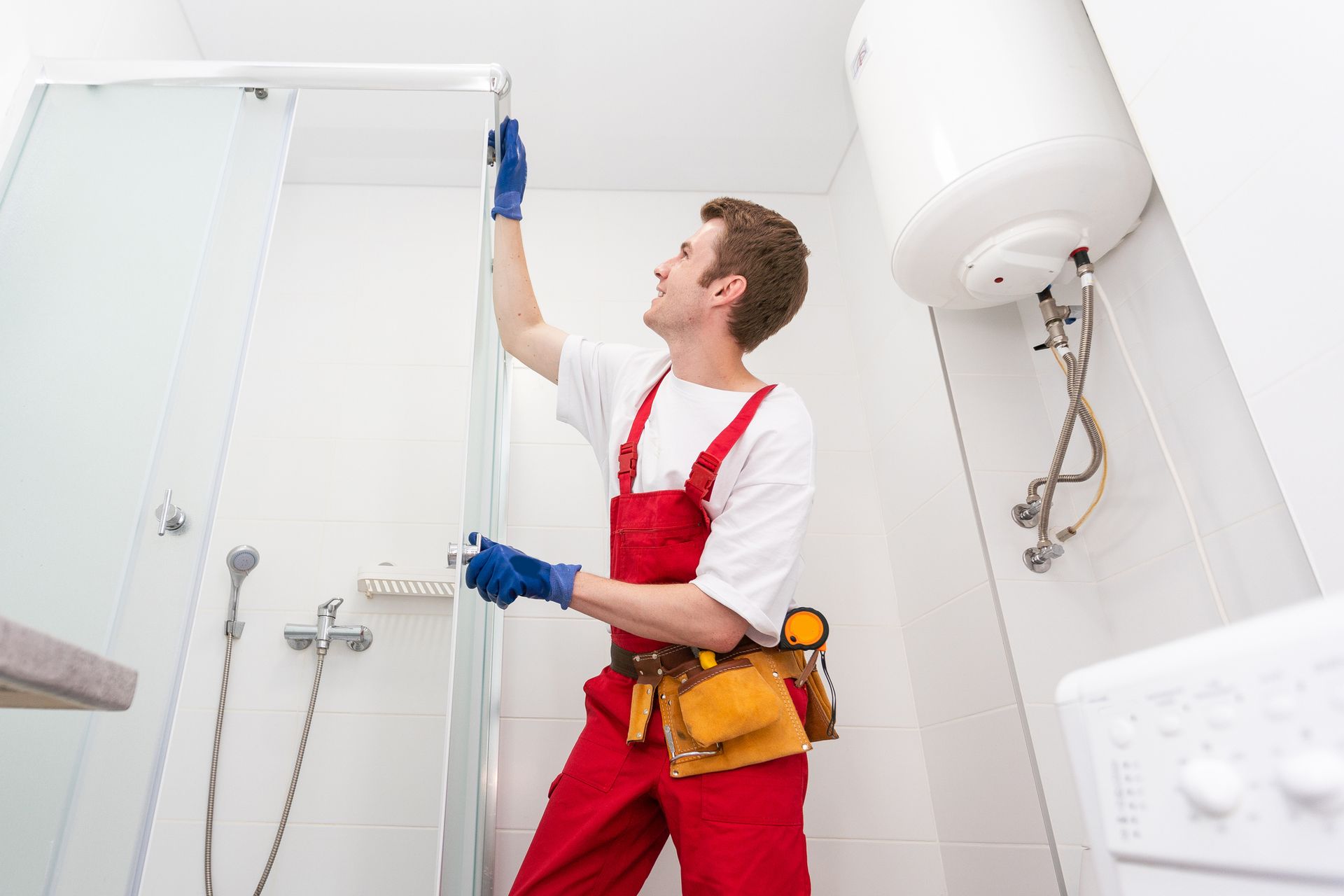 Man in red overalls installing a shower door in a bathroom, near a water heater.