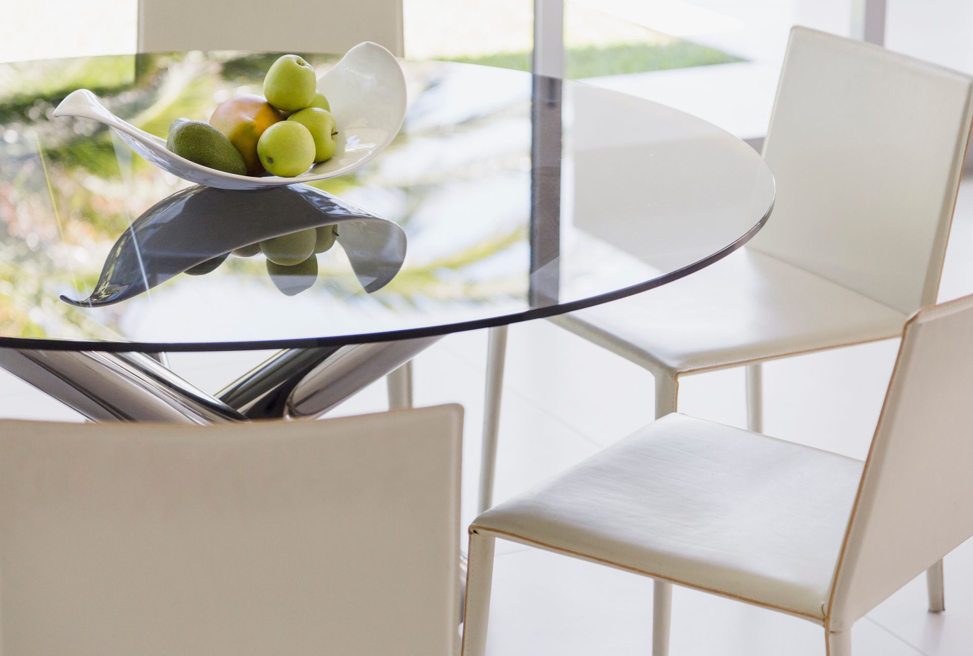 Round glass table with white chairs and fruit bowl.