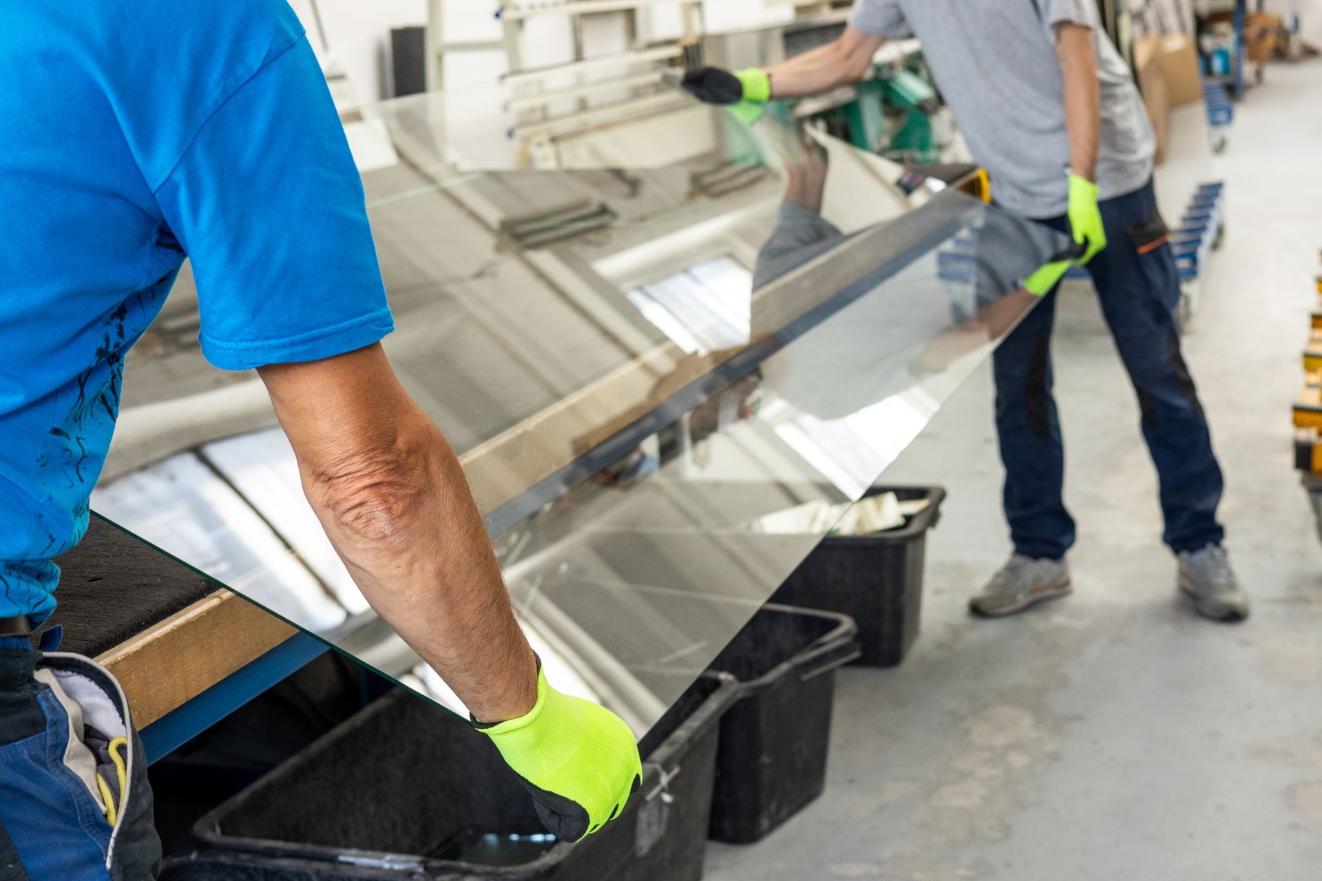 Two workers wearing gloves carefully handling a large sheet of glass in a factory setting.