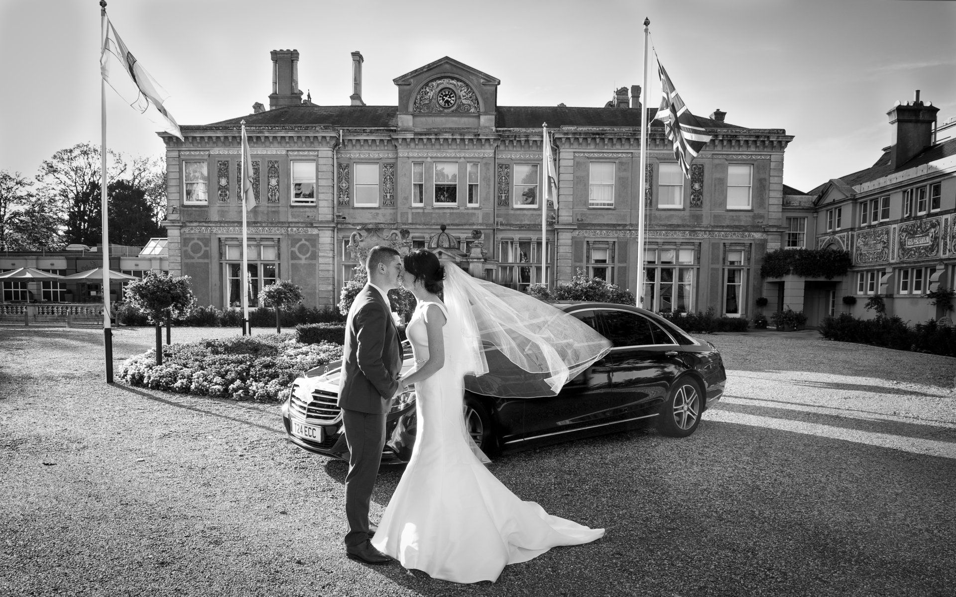 black and white portrait of bride and groom standing in front of a black Mercedes in the entrance to Down Hall hotel with flags fluttering in the wind