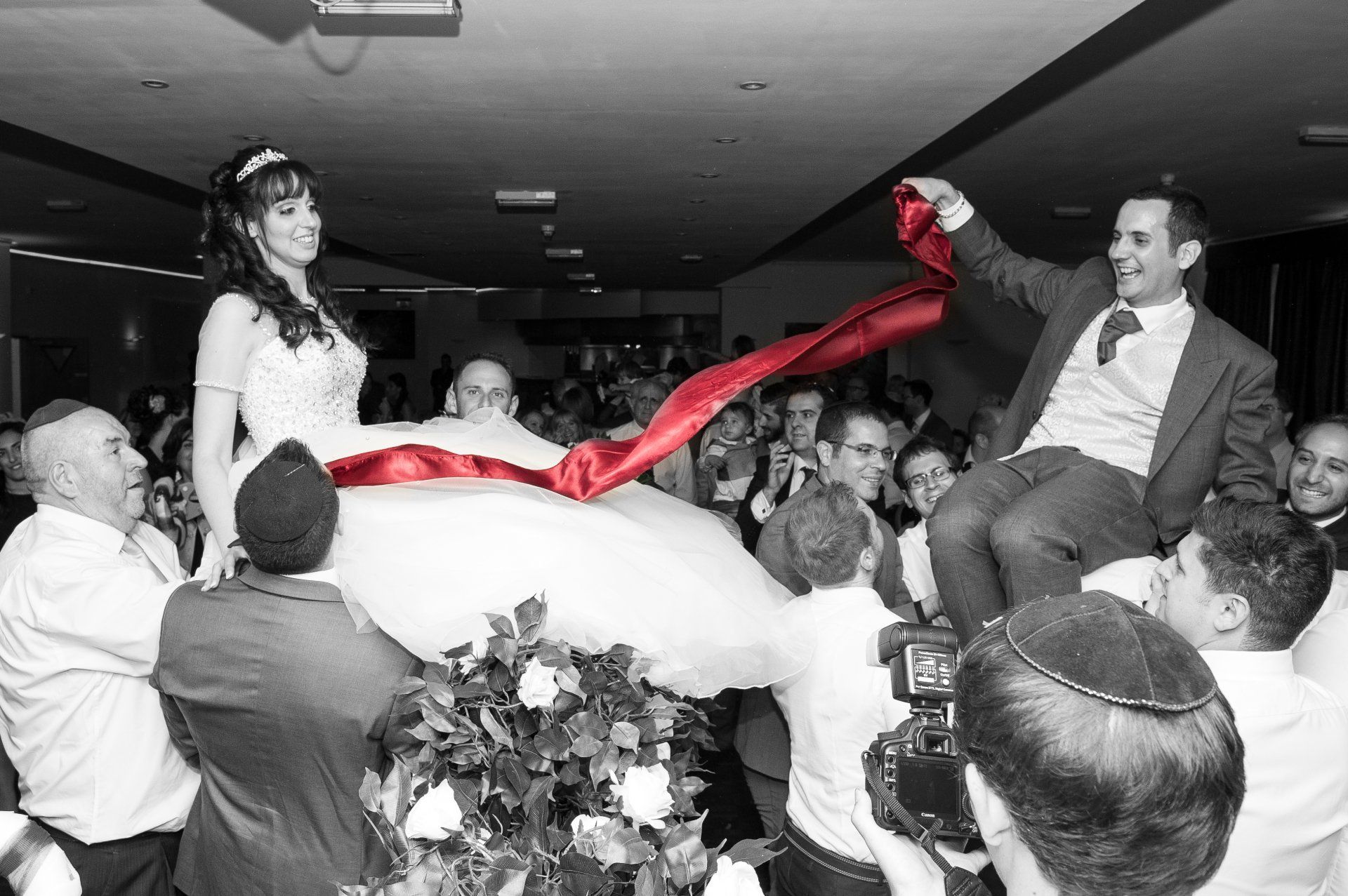 Jewish Bride and Groom sat on chairs, lifted into the air holding a red ribbon