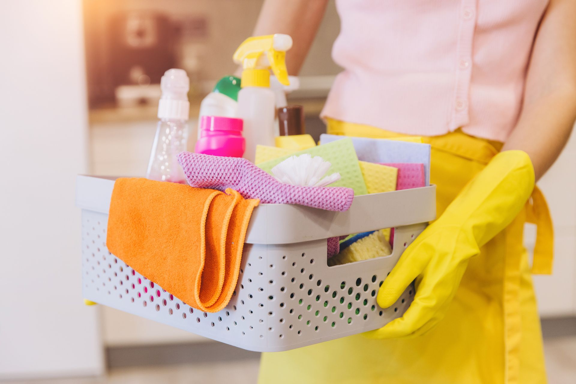 Woman wearing yellow rubber gloves holding basket containing cleaning products and equipment.
