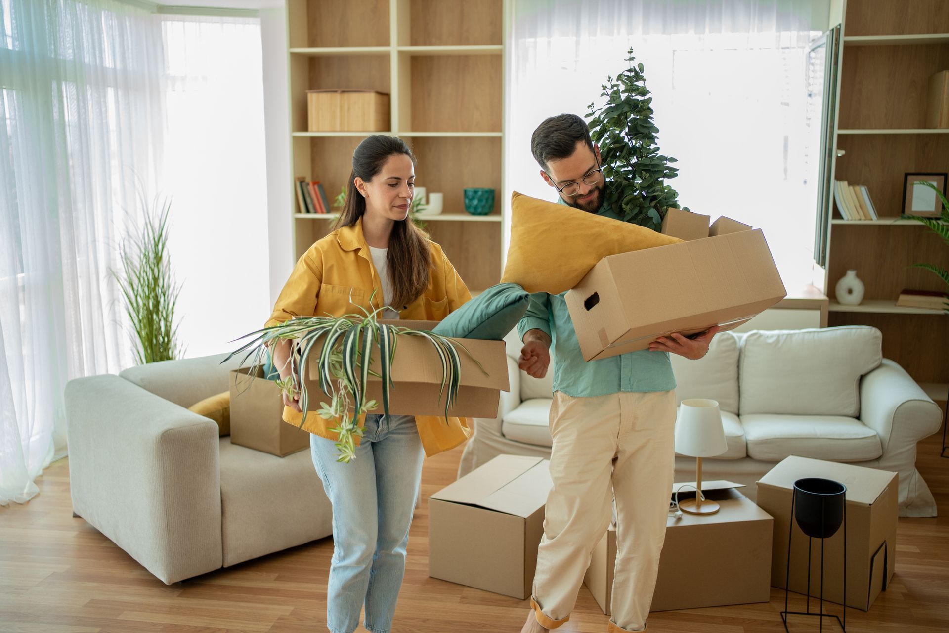 Couple moving boxes in a living room. Woman carries a box with plants, man holds one with a pillow and tree.