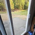 A sliding glass door open to a concrete patio and grassy yard with trees in the background.