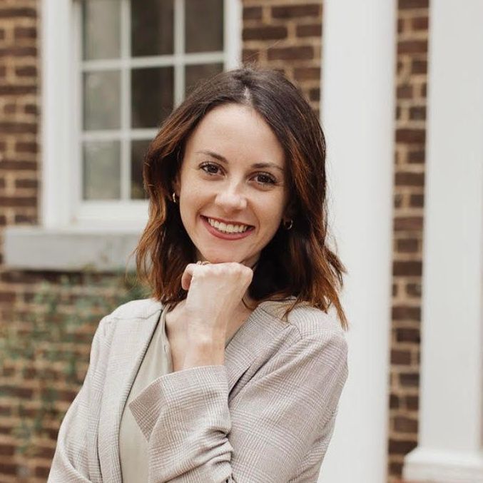 A woman is smiling with her hand on her chin in front of a brick building.