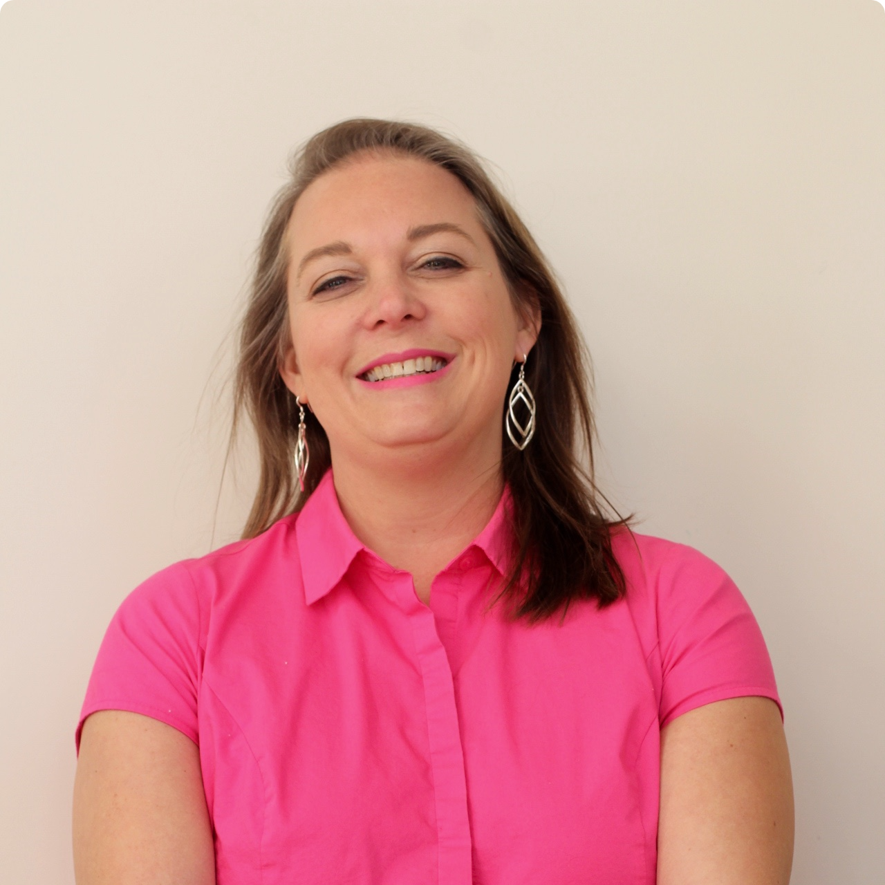 A woman wearing a pink shirt and earrings smiles for the camera