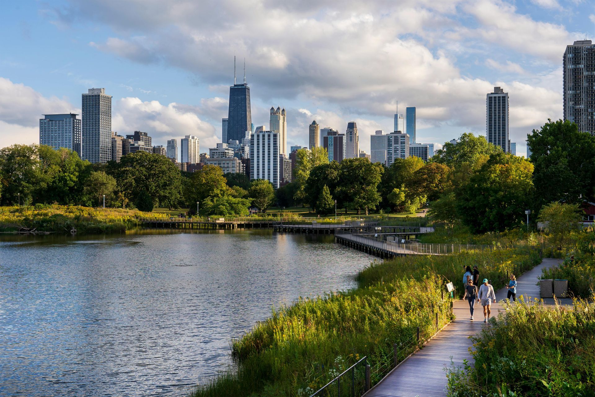 A group of people are walking along a path next to a lake with a city skyline in the background.