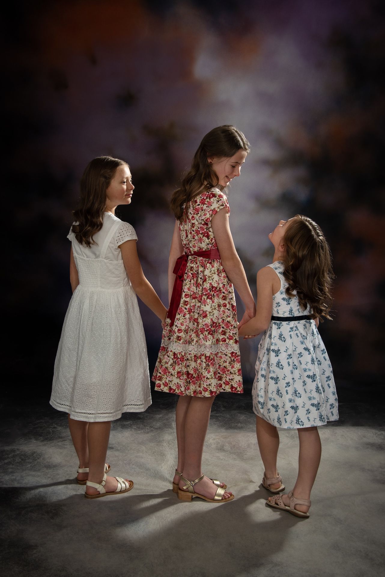 Three young girls in dresses hold hands, looking at each other in front of a cloudy background  — Dale Wright Portrait Maker in Shepparton, VIC