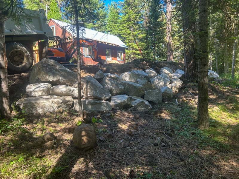 Large rocks form a retaining wall in front of a red building in a wooded area; a large machine is partially visible.