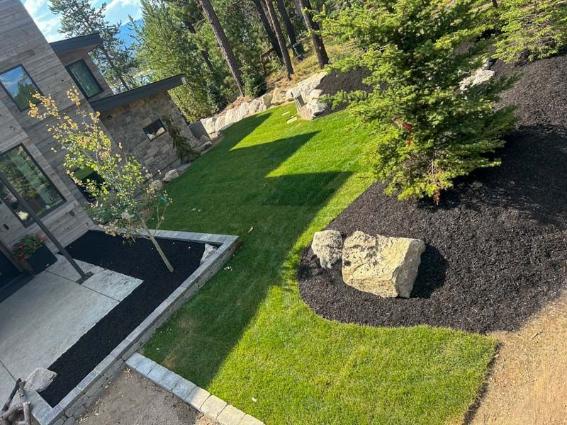 Lawn and landscaping around a modern house with a stone exterior. Black mulch beds contrast green grass.