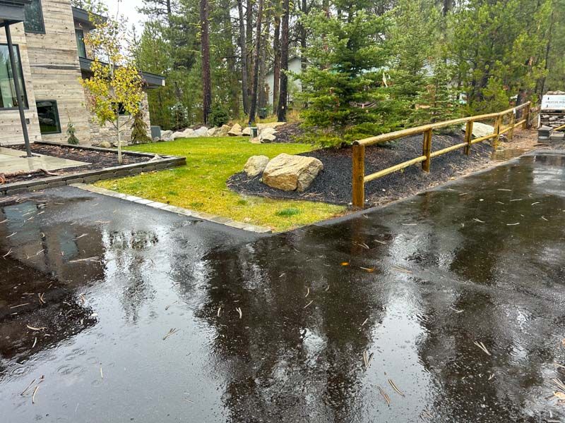 Wet asphalt driveway next to a small green lawn bordered by a wooden fence; trees and house in the background.