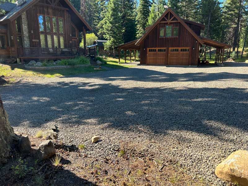 Gravel driveway leading to wooden house and garage in a forest setting with tall trees.
