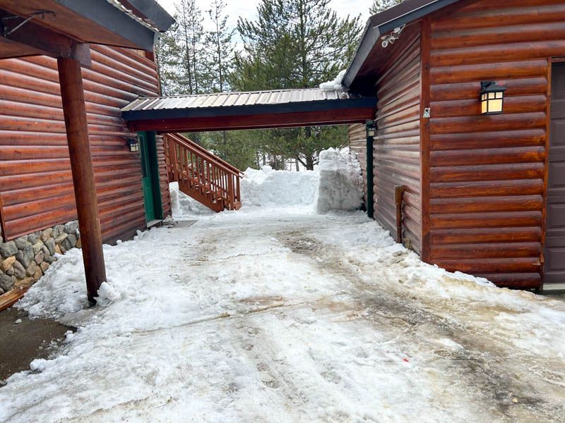 Driveway covered in snow, connecting two log buildings with a covered walkway above.