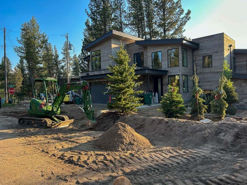 A green excavator in front of a modern house under construction. Brown dirt, young trees, and blue sky.