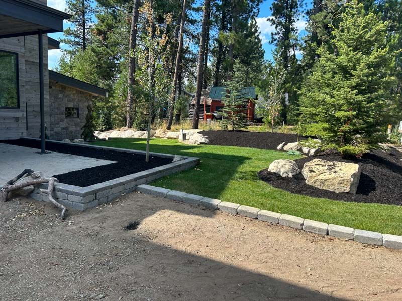 Landscaped yard with stone walls, gravel path, and green lawn, with trees and a cabin in the background.