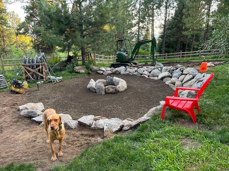 A fire pit under construction with a dog, rocks, and a green excavator in a backyard. Red chair.