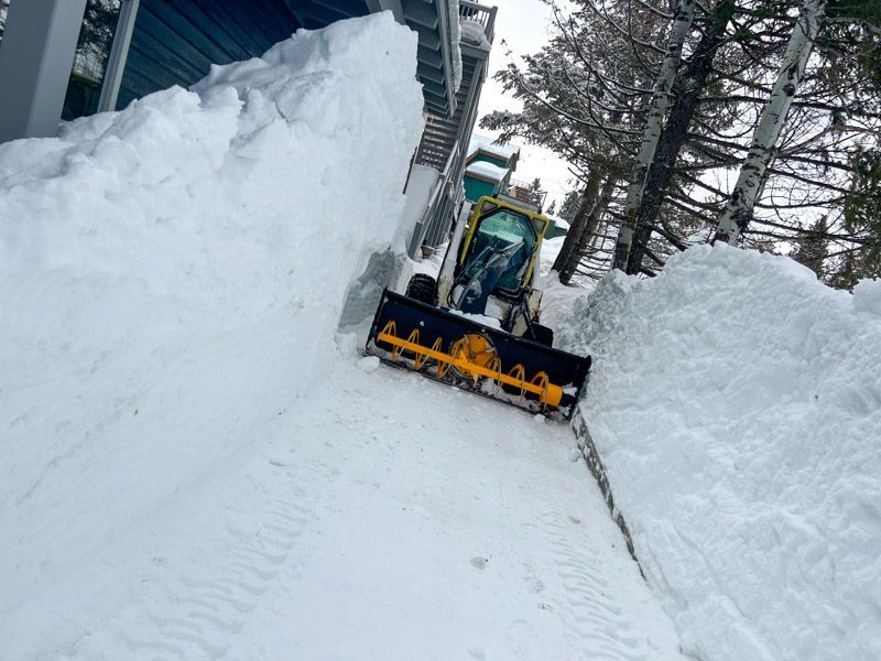 Small snowplow clearing a narrow path next to a building, high snow banks on either side.