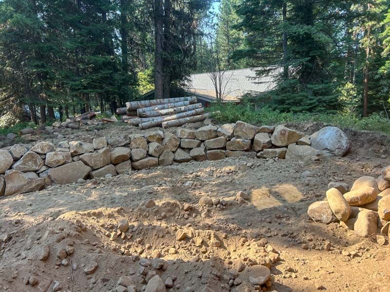 Construction site in a forest with a rock wall, dirt, logs, and a building under construction.