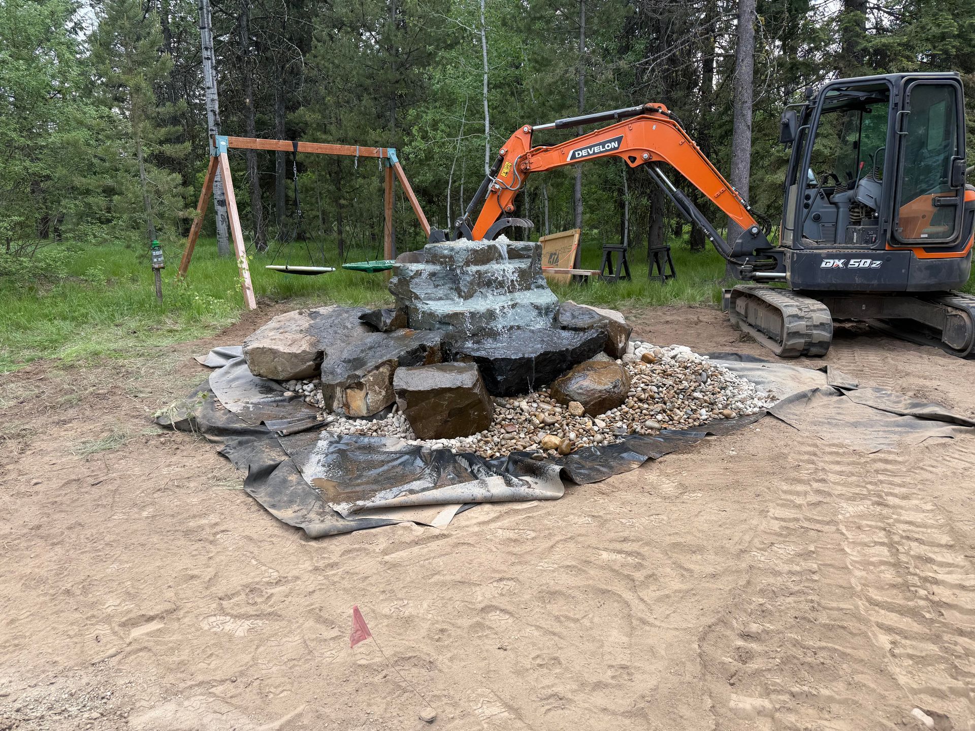An orange excavator placing large rocks to build a wall in a forest setting.