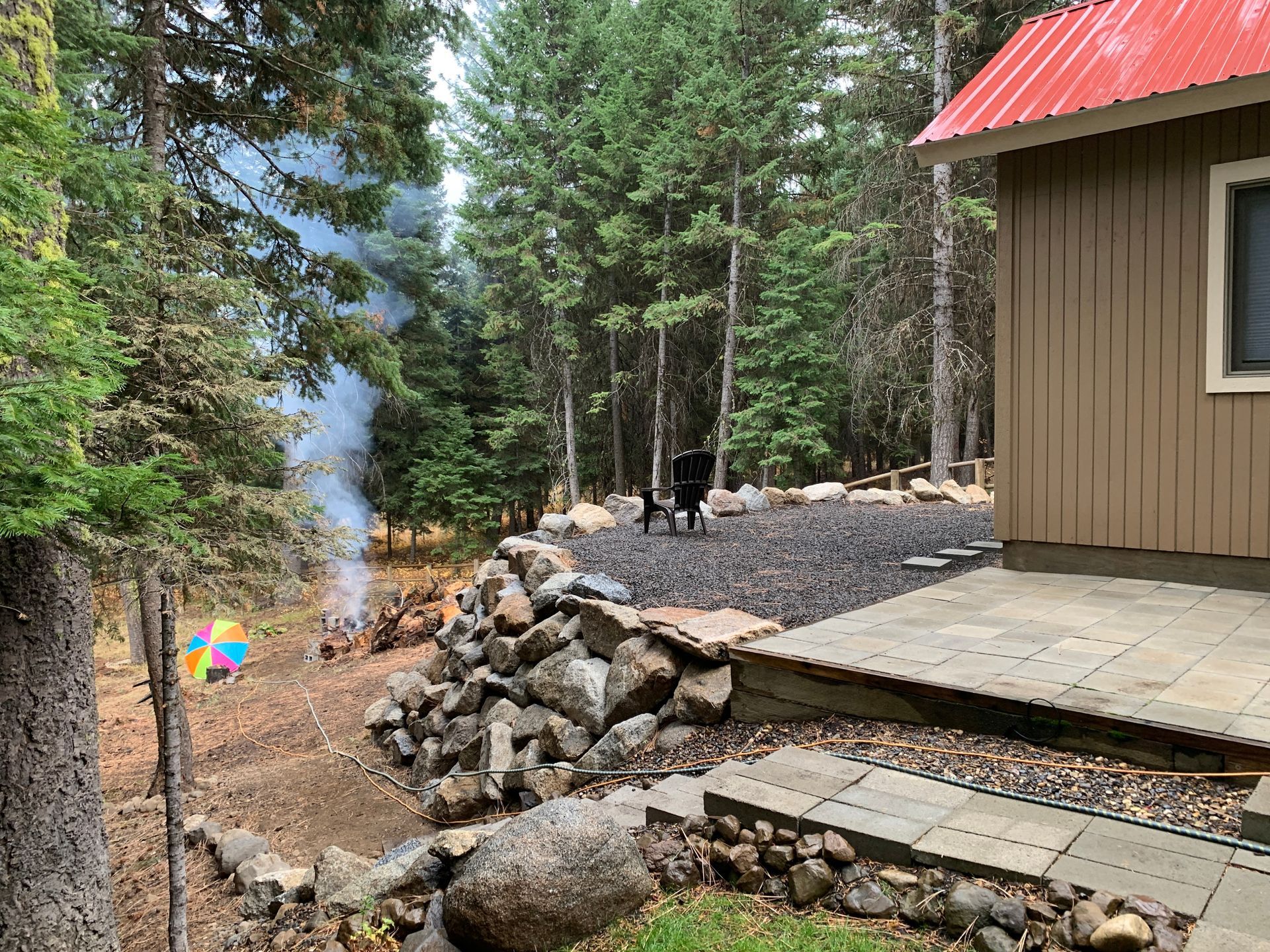 Smoldering campfire in a forested backyard; dog watches, building with red roof on right.