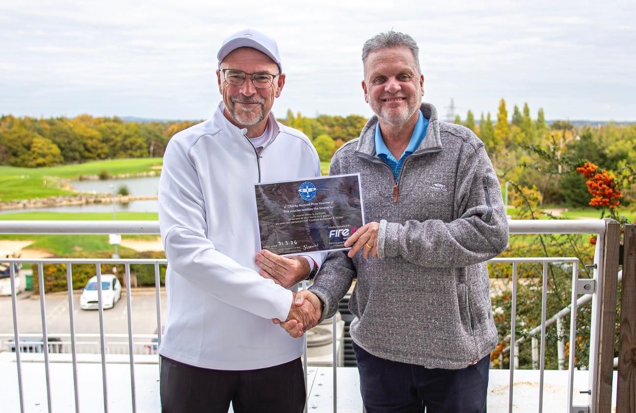Two men shake hands on a balcony, one holding a framed picture. Golf course visible in the background.