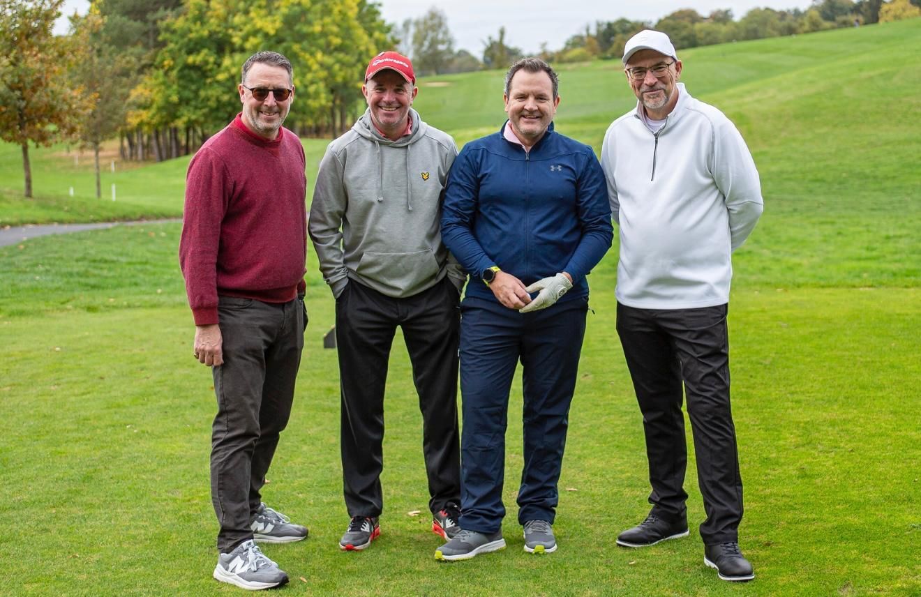 Four men stand on a golf course. They are wearing casual clothing. The ground is green with trees in the background.