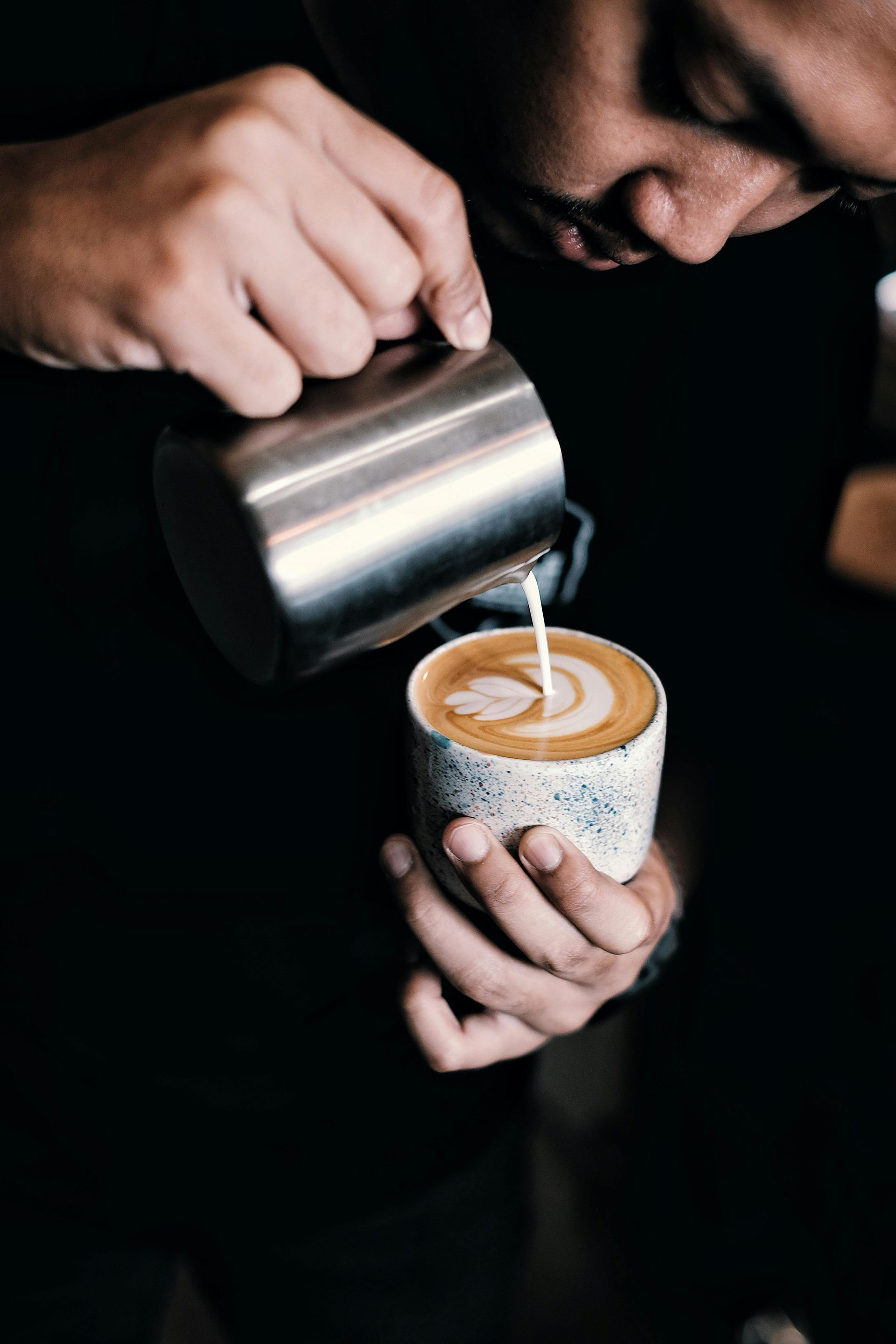 Barista pouring milk from a stainless steel pitcher into a cup of coffee, creating latte art.