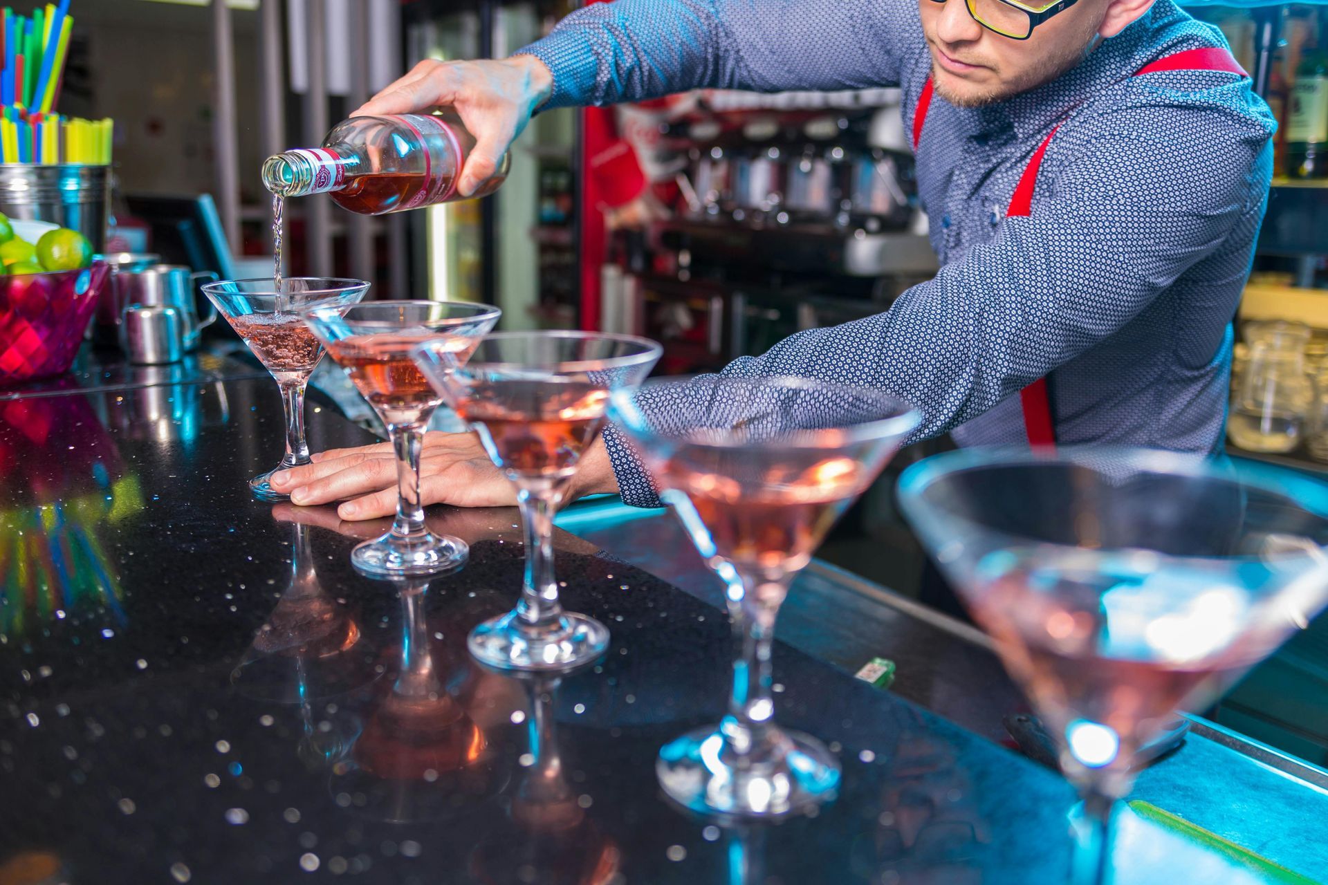 Bartender pouring liquid into martini glasses on a black, speckled bar.